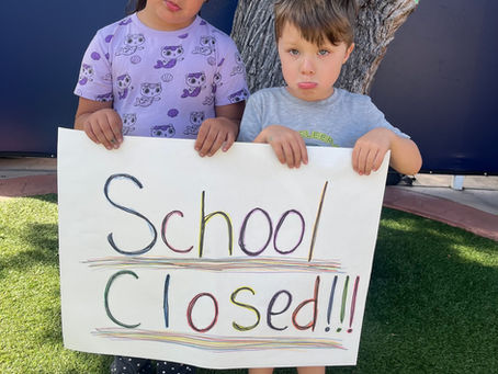Two young children stand outdoors in front of a tree, holding a handmade sign that reads “School Closed!!!” in colorful letters. Both children have sad expressions, and the sign is decorated with rainbow-colored outlines and lines.