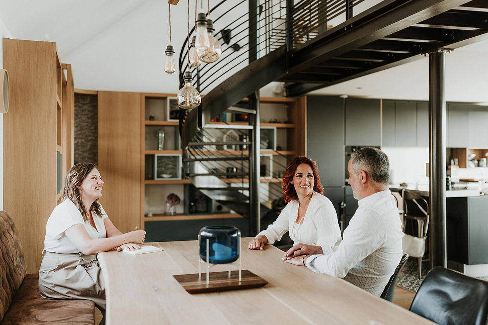 Trois personnes discutent souriantes autour d'une table en bois dans un salon moderne avec escalier en colimaçon et lampes suspendues. Un couple en rendez-vous avec son officiante pour préparer leur cérémonie laïque