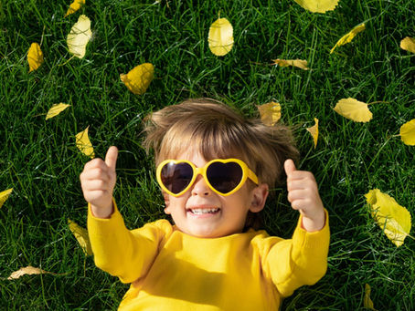 male toddler laying on grass with yellow heart-shaped sunglasses with 2 thumbs up