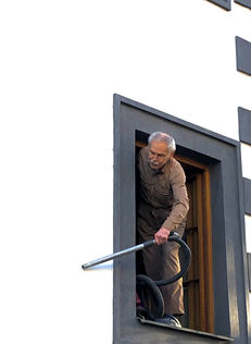 Picture of an older man vacumming the outside of a wall while standing inside the window frame of a building. 