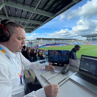 An AMS Audio Description commentator sits at his desk in the press area, speaking into a headset mic while checking his notes on the game in front of him.