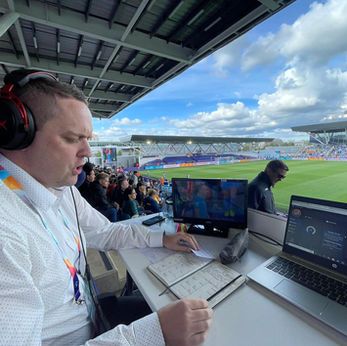 An AMS Audio Description commentator sits at his desk in the press area, speaking into a headset mic while checking his notes on the game in front of him.