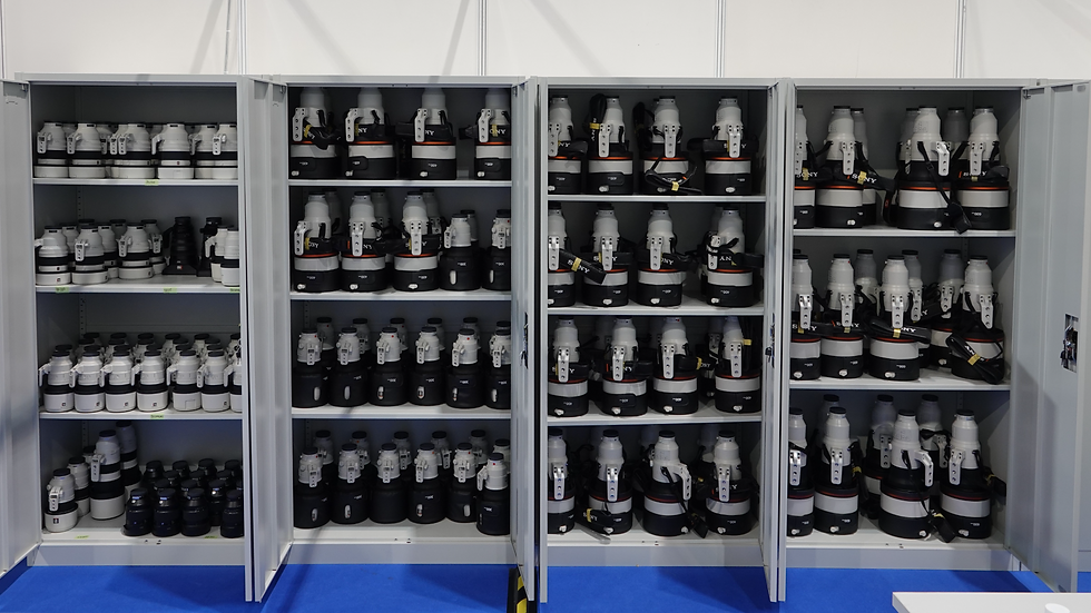 Shelves filled with white and black camera lenses in a storage room. The cabinets are open, revealing various lens sizes. Blue floor visible.