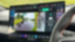 Car dashboard showing a rearview camera display with a wet road, greenery, and a bus. Steering wheel visible, text prompts for safety.