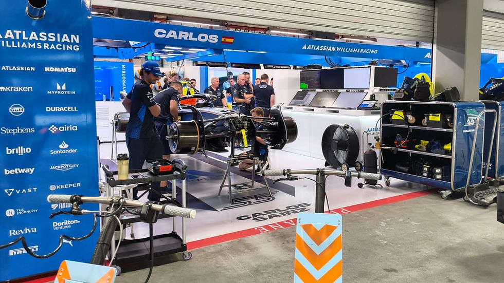 Mechanics work on a Formula 1 car in a pit garage. Blue Atlassian Williams Racing branding is visible. Tools and equipment surround the car.