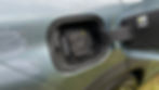 Car's charging port open on a blue vehicle with a Kumho tire. Reflection of a white shirt visible. Grass in the background. Warning sticker present.