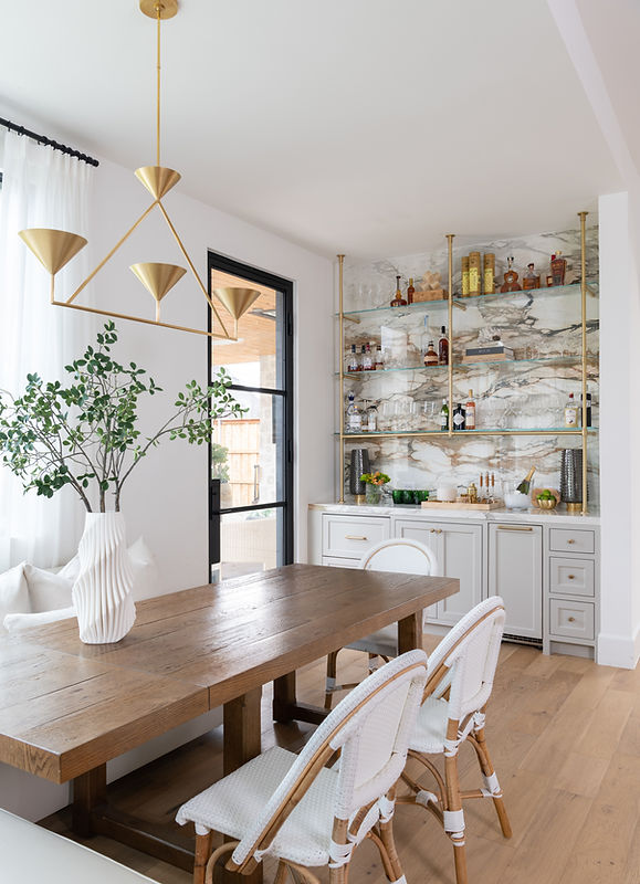 Modern mediterranean breakfast nook featuring a wooden dining table, white Serena and Lily chairs, a porcelain slab backsplash, and gold palmer shelving with glass shelves styled with bar accessories.