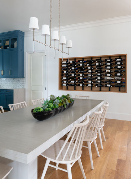 Transitional dining room design with white dining chairs, a long black onyx table arrangement, a silver and white light fixture, and a cased wine bottle wall rack.