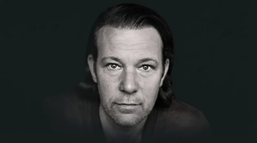 Studio portrait photography of a man with dark hair and a serious expression. The lighting is moody, highlighting his direct gaze.