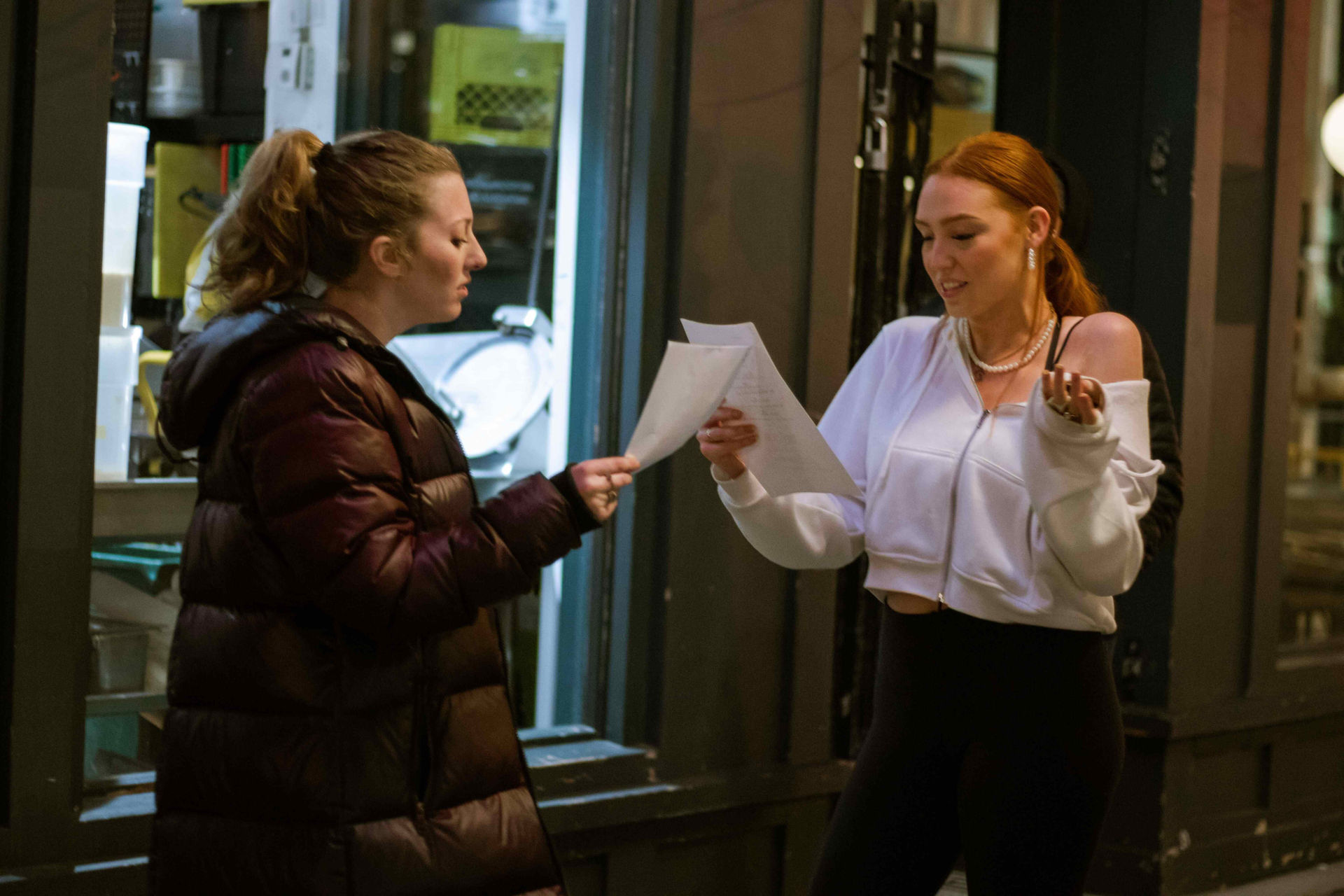 Two women review papers on street; woman gestures, sharing documents, night setting.