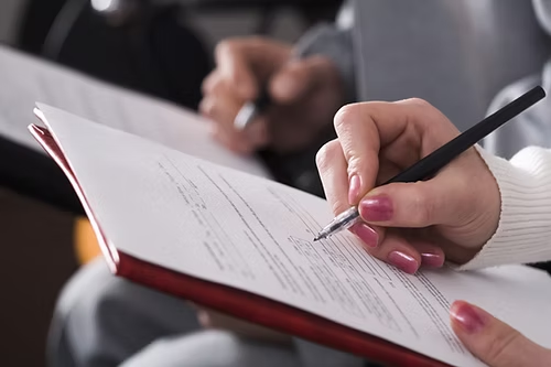 Person signing paperwork with pen, hands holding pen, red folder, document in process.
