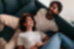 Young couple laid together laughing, dressed in light casual clothing with a teal sofa as the background.