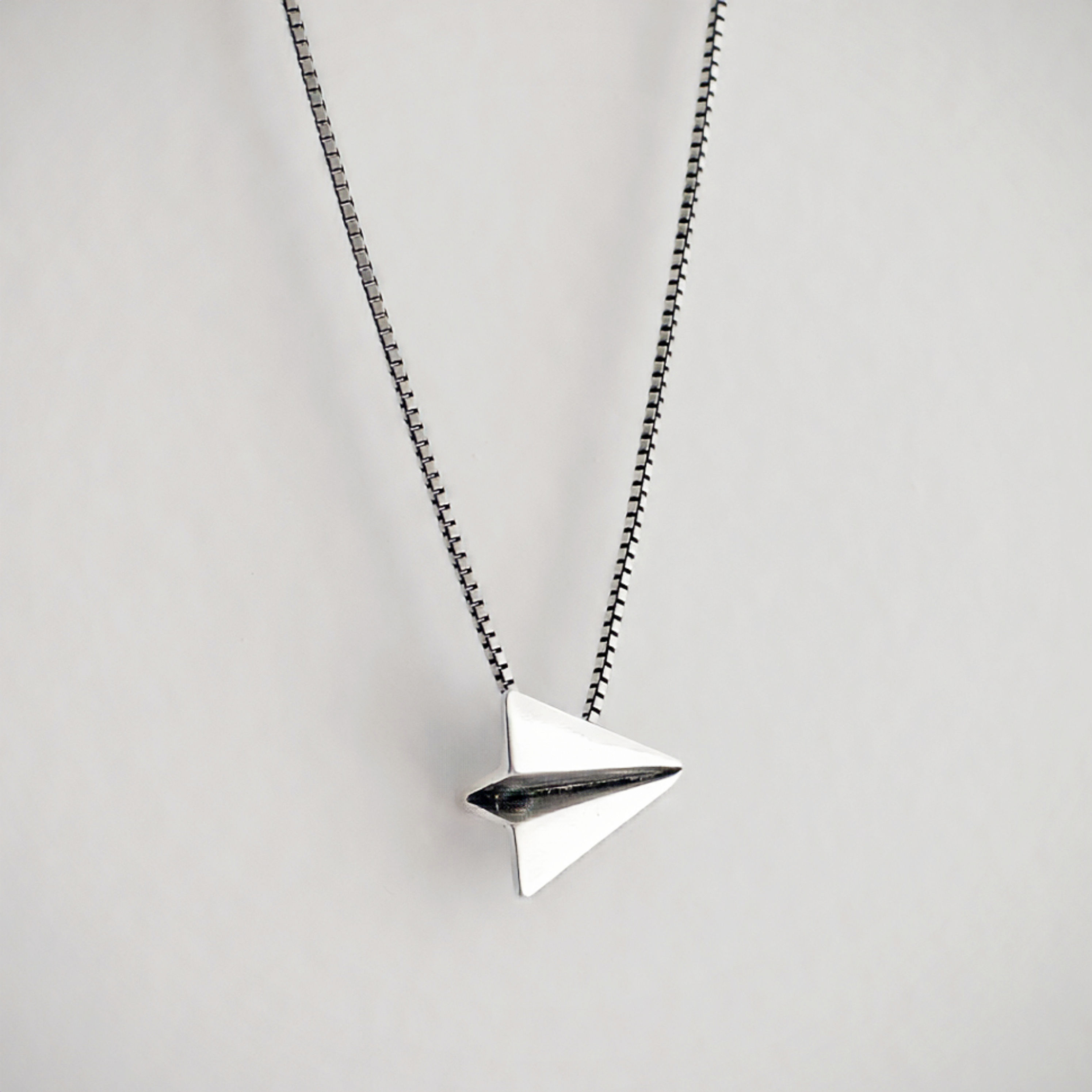 Colar Gaivota com cordão prateado e pingente em formato de aviãozinho de papel, também em prata.