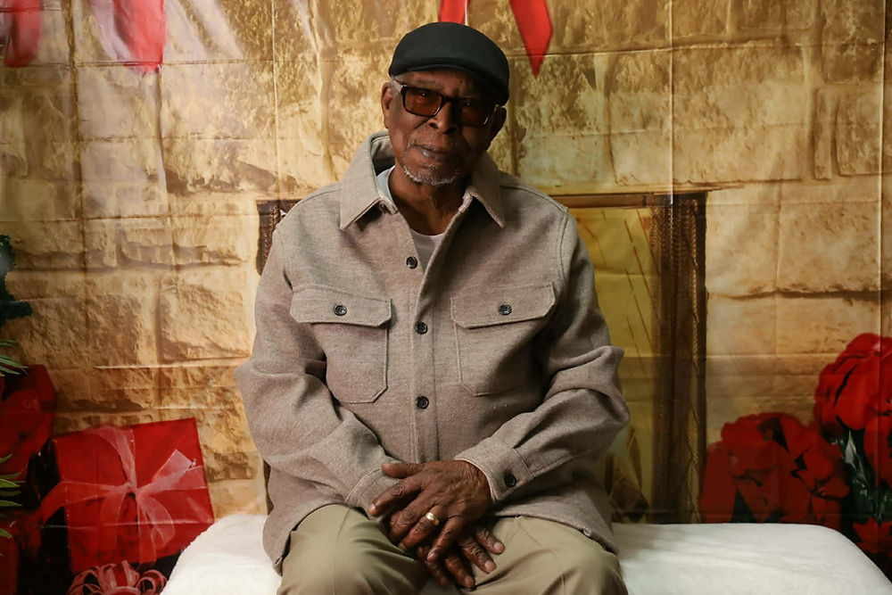 Older man in a jacket and cap, sitting against a festive backdrop.