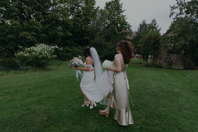 Bride walking across lush green lawn with bridesmaid holding her veil during an outdoor Vermont wedding at a New England wedding venue