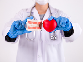 Doctor in blue gloves holds a dental model and a red heart shape. Wears white coat, stethoscope; neutral background.