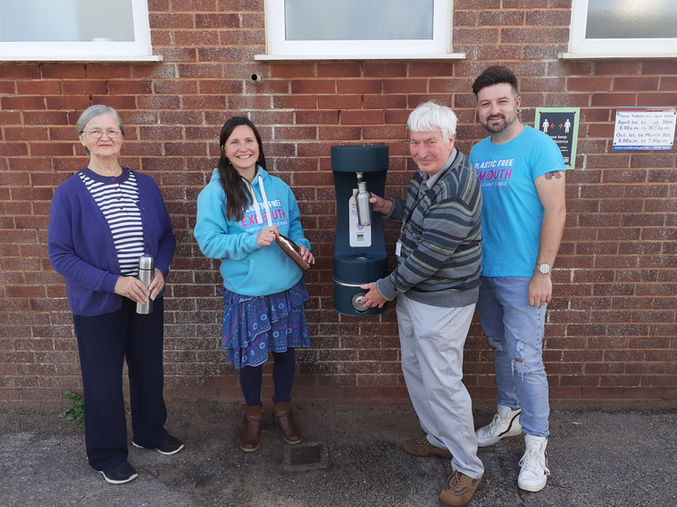 Four people stand near a water bottle refill fountain near beach