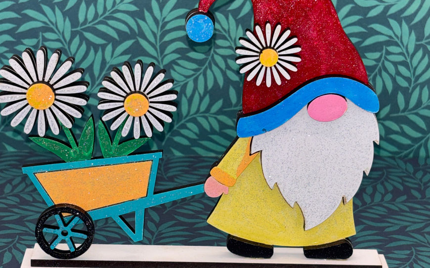 A 3D layered wooden craft figure of a walking gnome with a red glittery hat pushing a teal and orange wheelbarrow filled with white daisies.