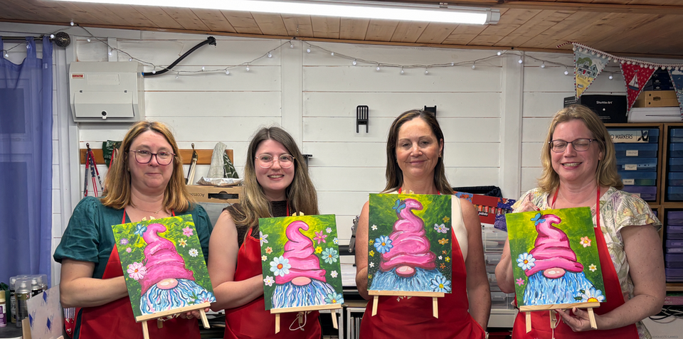 Four smiling women wearing red aprons proudly holding up their finished hand-painted summer garden gnome canvases inside a well-organised art studio workshop.