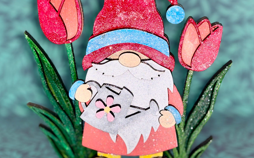 A freestanding layered wooden gnome craft wearing yellow boots and a red hat, holding a silver watering can while standing between two large red painted tulips.