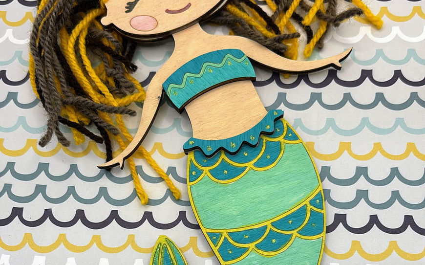 A laser-cut wooden mermaid figurine with flowing yellow and brown yarn hair, painted in a beautiful gradient of teal and emerald green ocean scales.