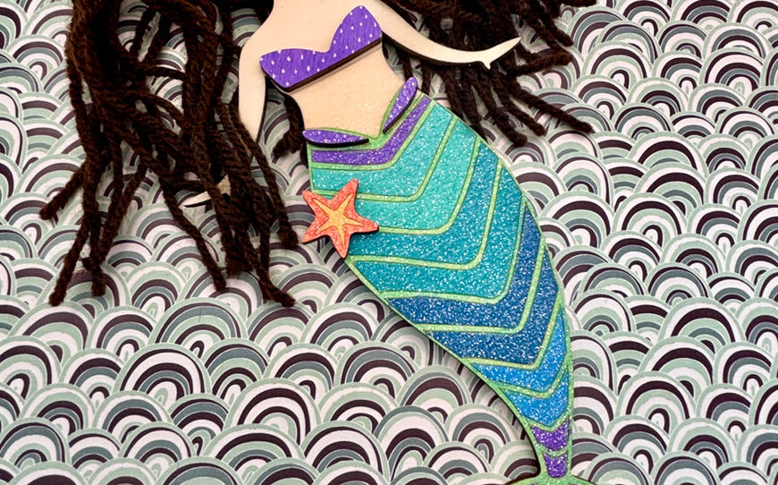 A wooden mermaid figurine with long, dark brown yarn hair that appears to float, holding an orange starfish on her glittery blue and green chevron-patterned tail.