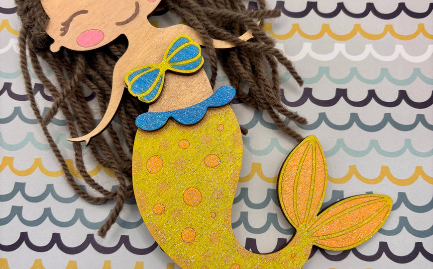 A whimsical wooden mermaid craft with textured brown yarn hair, a vibrant blue top, and a sparkling, glittery yellow and orange tail.
