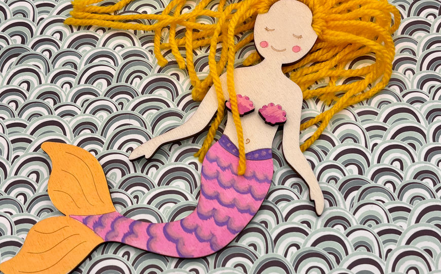 A handcrafted laser-cut wooden mermaid doll with bright yellow yarn hair and a hand-painted pink and purple scaled tail, resting against a wavy patterned background.