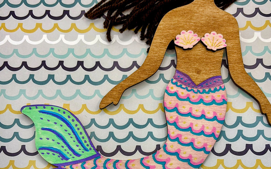A wooden mermaid craft figure featuring dark brown yarn hair and a detailed pink, purple, and green hand-painted tail with glitter highlights.