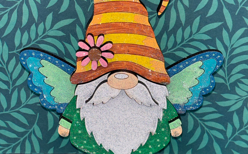 A freestanding 3D wooden gnome figurine painted with green robes, a silver glittery beard, an orange striped hat, and sparkling blue-green butterfly wings.
