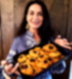 Woman smiling, holding a tray of pastries. She wears a blue embroidered shirt and colorful earrings. Wooden background.