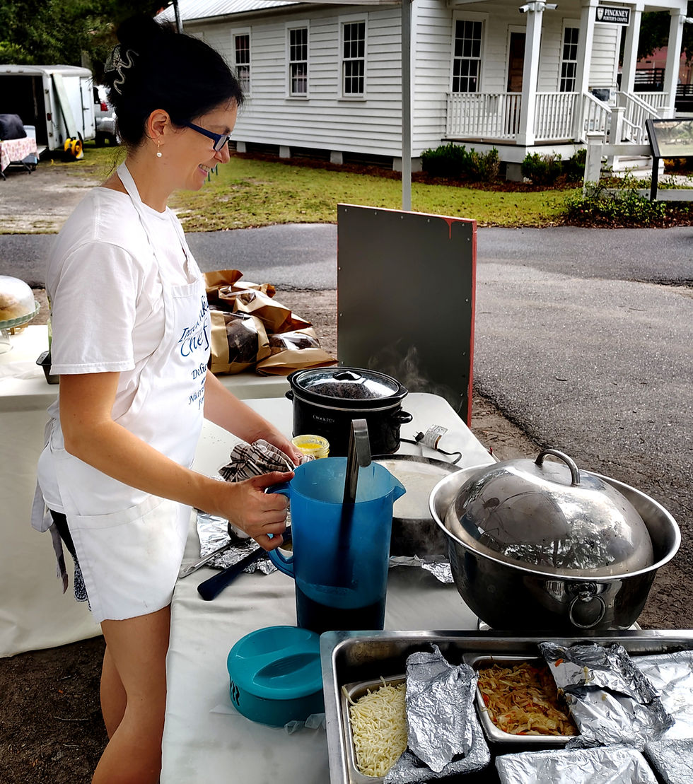 Woman in apron serves food at an outdoor table with steaming pots and trays, beside a white house with a Finckney sign.
