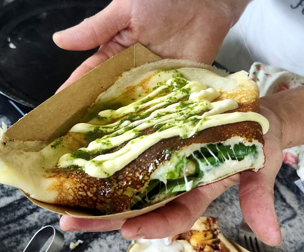 Hands hold a stuffed crepe in a paper tray, topped with white sauce and green drizzle, in a kitchen close-up.