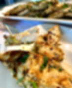 Grilled chicken fillet on a fork, garnished with herbs. More fillets on a platter in the background, set on a white table. Appealing and fresh.