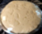 Risen dough covered with plastic wrap in a metallic bowl on a dark countertop. The dough appears airy with visible texture.