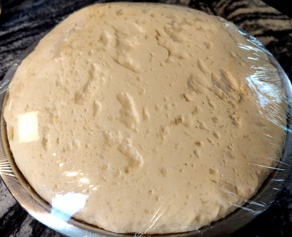 Risen dough covered with plastic wrap in a metallic bowl on a dark countertop. The dough appears airy with visible texture.