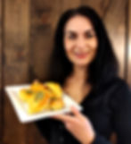 Woman smiling, holding a plate of baked pastries with green filling. Wooden background adds a rustic feel. Mood is warm and welcoming.