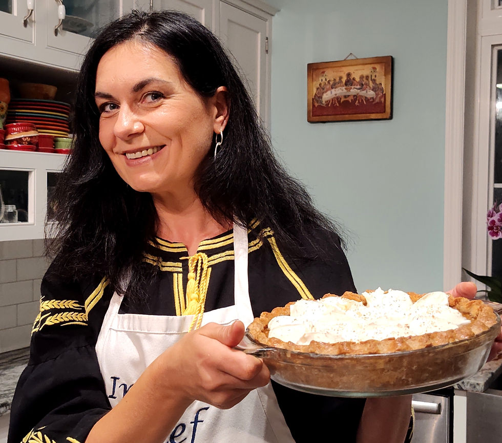 Smiling woman in a kitchen holds a cream-topped pie. She's wearing an apron and a black shirt with gold accents. Shelves and art are visible.