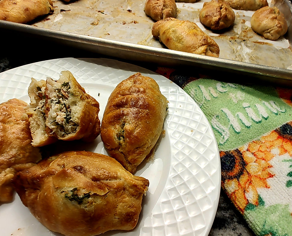 Baked pastries on a white plate, one cut open showing filling. Background features tray with more pastries and a colorful towel.