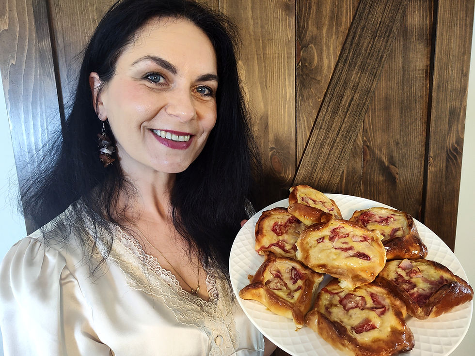 Woman smiling, holding a plate of pastries with raspberry filling. She wears a cream blouse and earrings. Wooden backdrop.