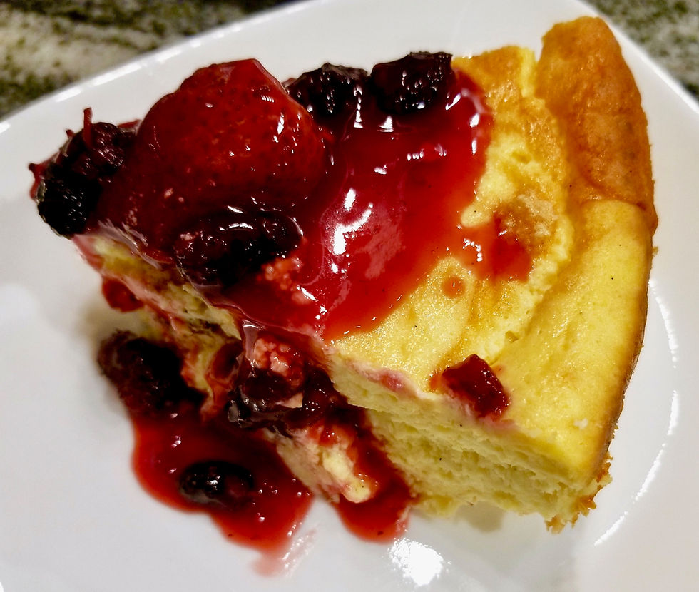 A slice of cheesecake topped with glossy red strawberry sauce and berries on a white plate. Soft texture contrasts with vibrant topping.