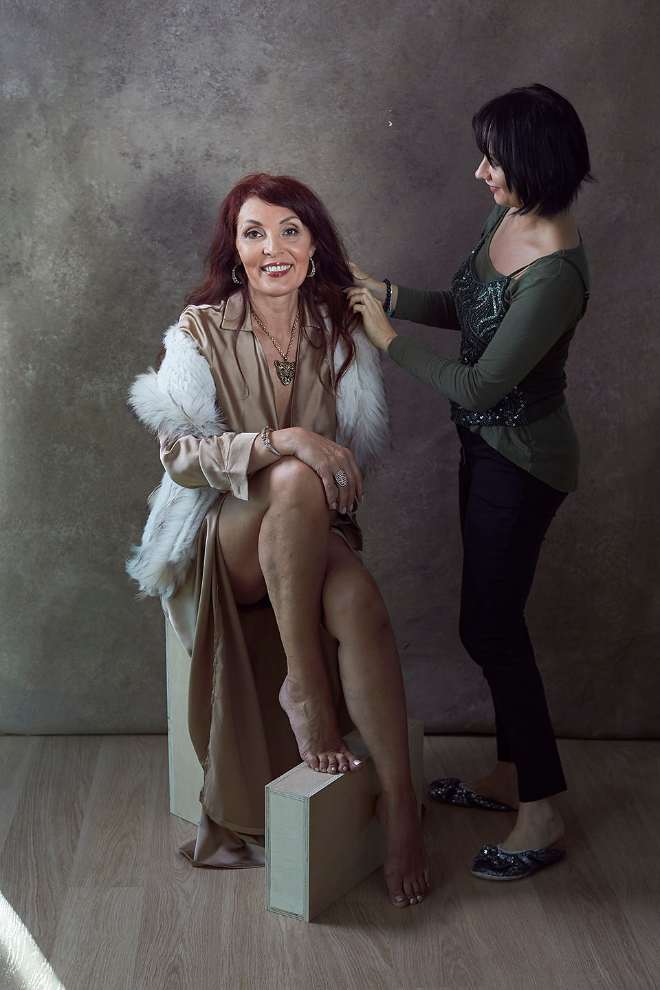 Long dark red hair woman wearing white sleeveless fur jacket on top of golden beige silk long dress, sitting opposite beige studio Gravity background while a short dark hair makeup artist wearing khaki color top and dark jeans touching up her hair before the 45 Over 45 Project photoshoot.