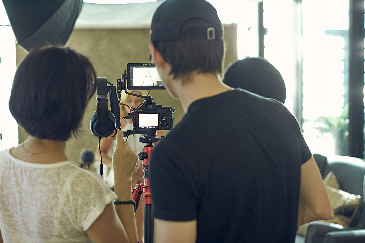 Two backs of a young video producer wearing black t-shirt and baseball hat, and a dark short hair lady-interviewer wearing white t-shirt, checking the back of the camera before recording their interview for the 45 Over 45 Project participant.