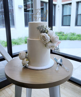 Elegant two-tier white wedding cake decorated with white peonies and greenery, topped with a custom gold topper reading 'The Fosters,' displayed on a wooden table by large glass windows with a cake knife and server set beside it.