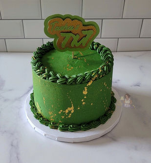 An apple green colored cake with the same colored buttercream borders around the top and bottom of the cake. There is gold splatter on the cake. There is a custom cake topper  in the same colors of the cake with "Happy 7X7" on it.