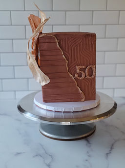 Elegant square cake finished in mauve buttercream with textured geometric patterns, metallic gold accents, and the number '50' displayed on the side. Adorned with striking edible gold wafer paper sails, showcased on a metal cake stand against a white tiled background.