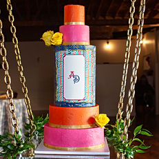 A Carnivale themed wedding cake, 5 tiers tall, the top and 4th tier are bright orange, the 2nd and 5th tier are bright pink, they all have gold ribbon around the bottom. The 3rd (middle) tier is a deep blue with a logo to match the wedding invitations. There are 3 gold roses placed around the cake. The cake is on a platform suspended in air with gold chains and surrounded by greenery