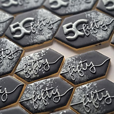 an array of elegantly decorated 50th birthday custom cookies, black with silver touches. some have the number "50" with the word "fifty" written through the "0" and others with the word "fifty" written across the front