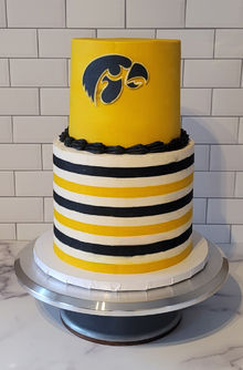 2 tiered cake in mustard yellow, black, and white. Bottom tier with horizontal stripes in mustard yellow, black, and white the top tier in mustard yellow with the Hawkeye logo in black on front with a black buttercream boarder at the base.