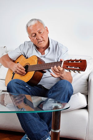 Older man playing acoustic guitar on a white sofa in a relaxed casual home setting with glass coffee table in foreground for Kingwood Arts Academy blog..